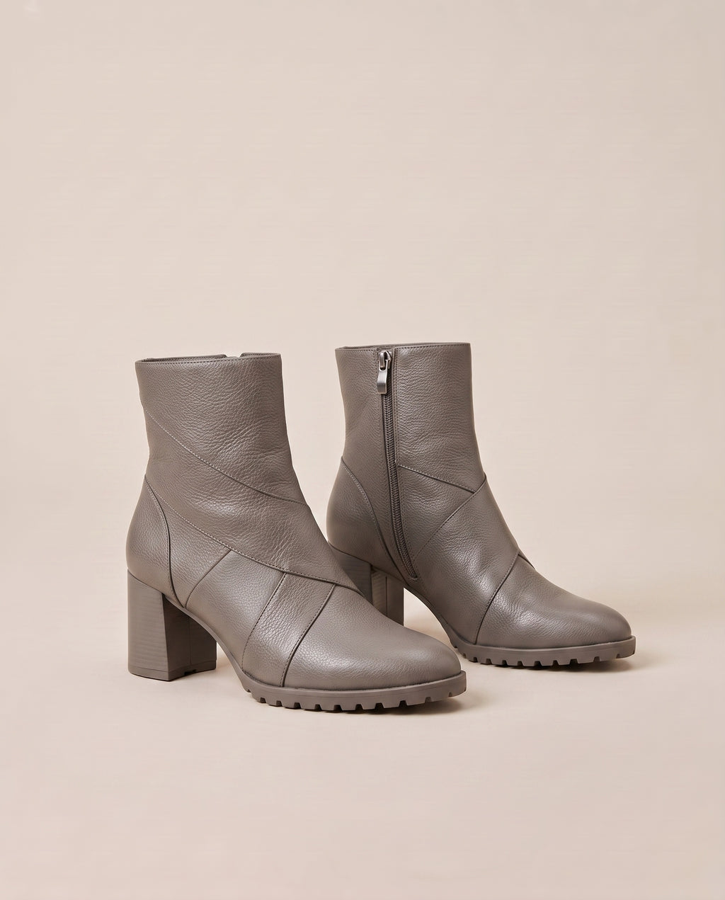 A pair of taupe gray ankle boots with block heels, side zippers, and chunky lug soles, photographed on a solid beige background.