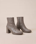 A pair of taupe gray ankle boots with block heels, side zippers, and chunky lug soles, photographed on a solid beige background.