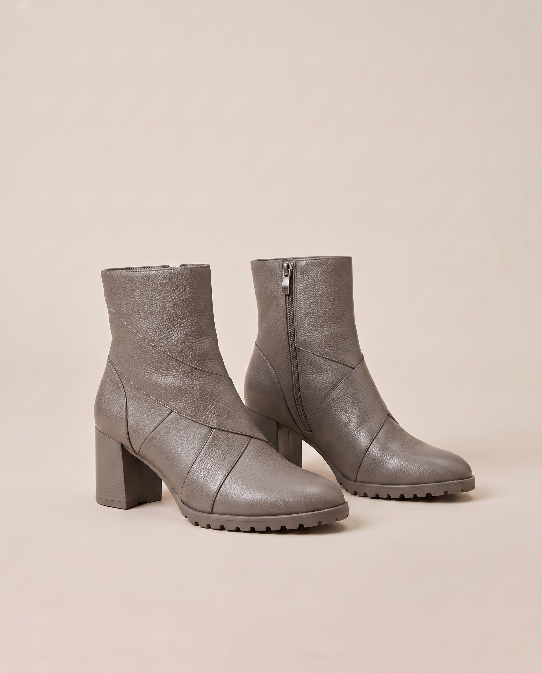 A pair of taupe gray ankle boots with block heels, side zippers, and chunky lug soles, photographed on a solid beige background.