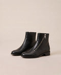 A pair of black ankle boots with a low block heel and a silver zipper on the side, displayed against a solid beige background.