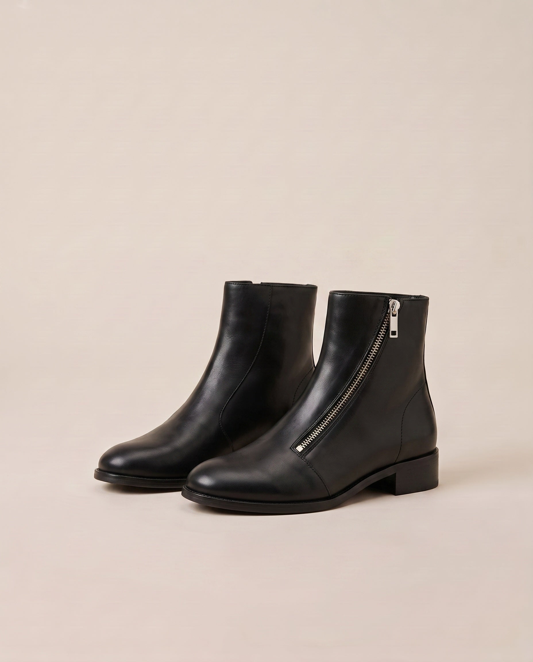 A pair of black ankle boots with a low block heel and a silver zipper on the side, displayed against a solid beige background.