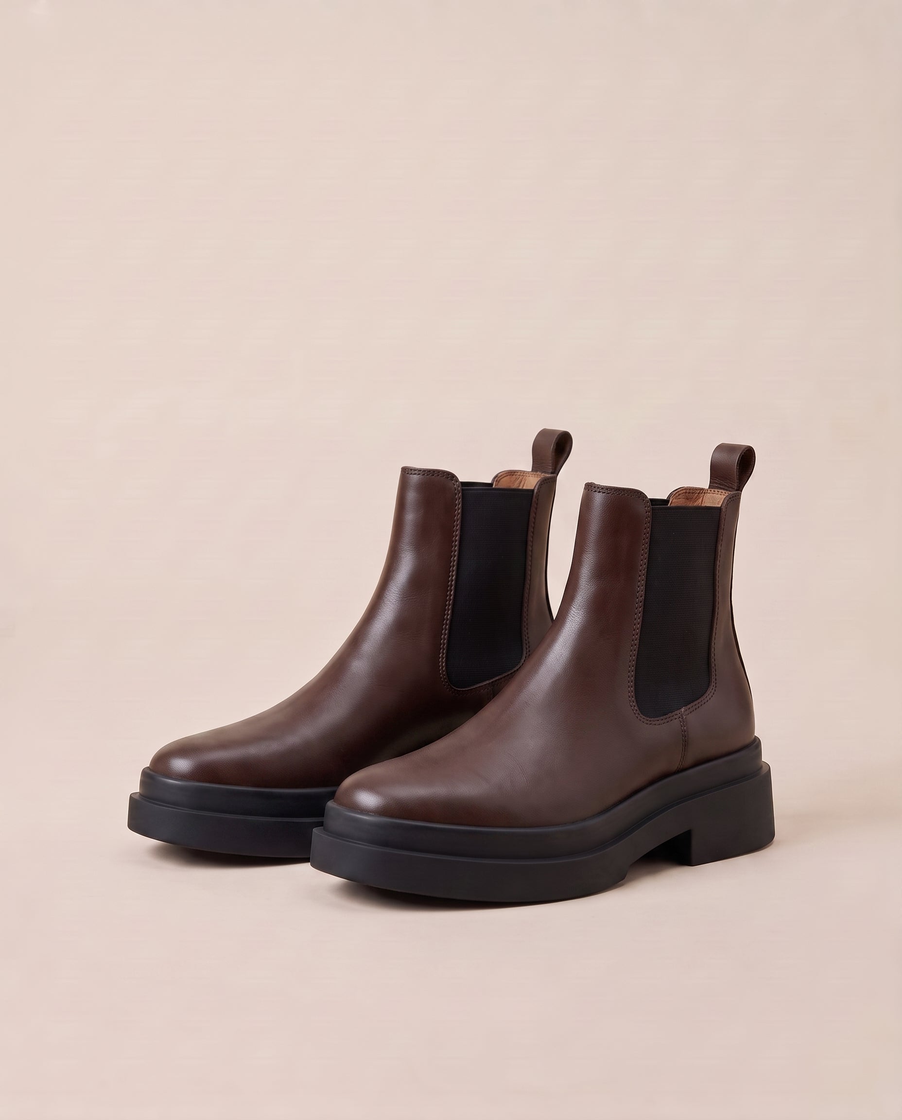 A pair of dark brown Chelsea boots with black elastic side panels and chunky black platform soles, displayed on a warm beige background.