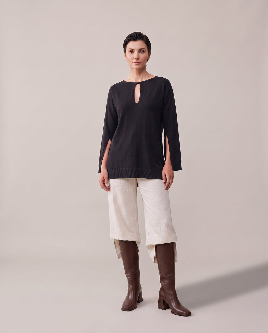 A woman stands against a plain light beige studio background wearing a black long-sleeve top with a small keyhole cutout at the neckline, off-white cropped pants, and brown knee-high boots.