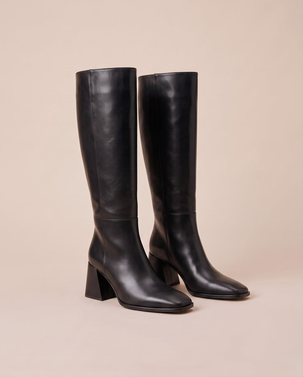 A pair of black knee-high boots with block heels and square toes, displayed on a solid beige background.