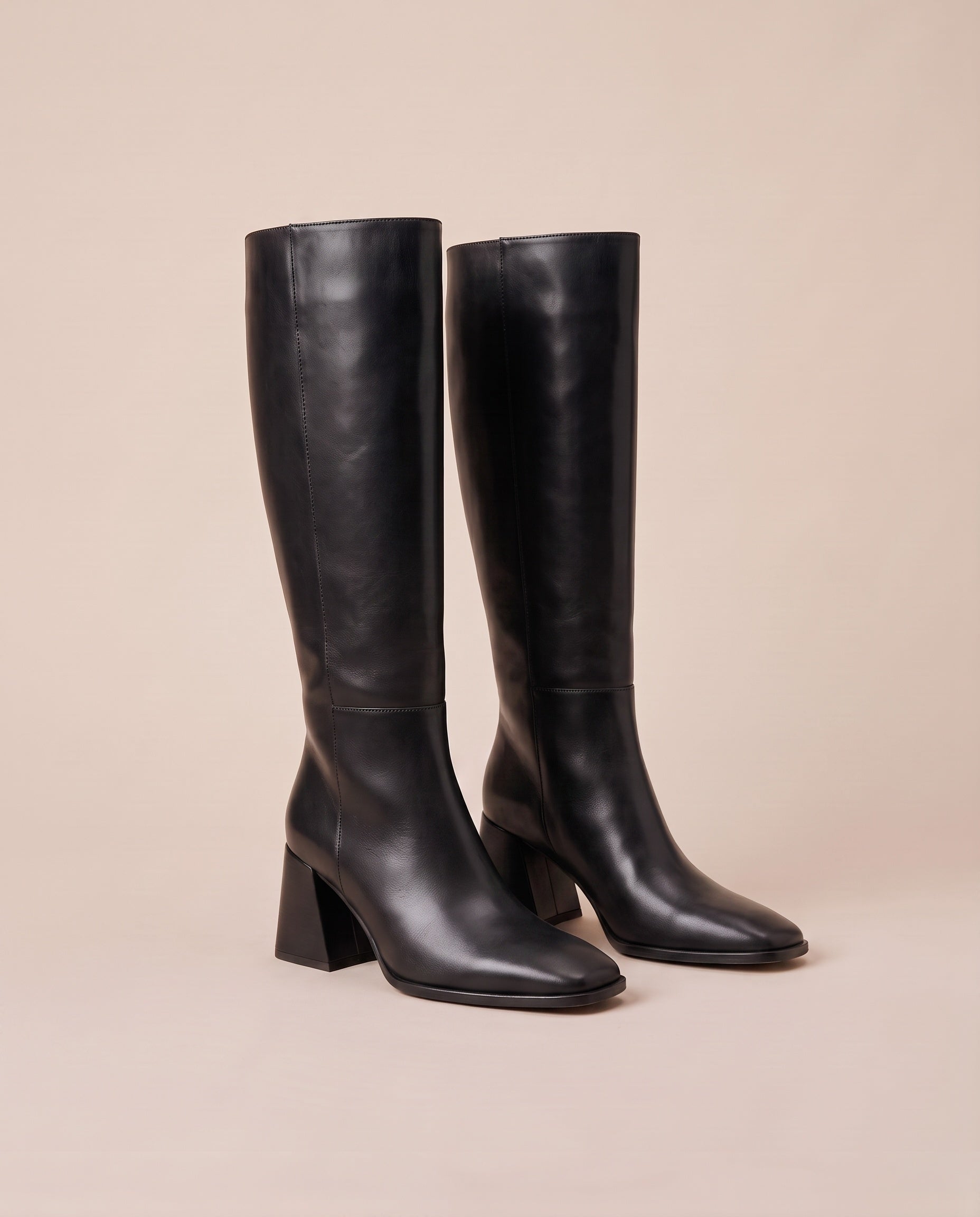 A pair of black knee-high boots with block heels and square toes, displayed on a solid beige background.