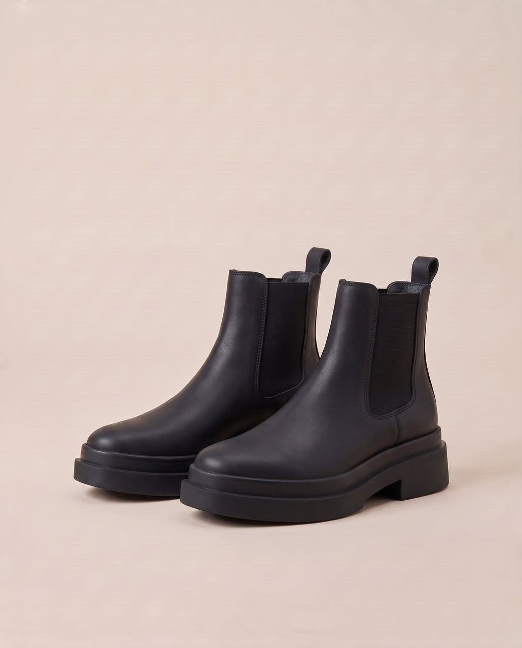 A pair of black Chelsea boots with chunky platform soles, shown from a three-quarters angle on a solid beige background.
