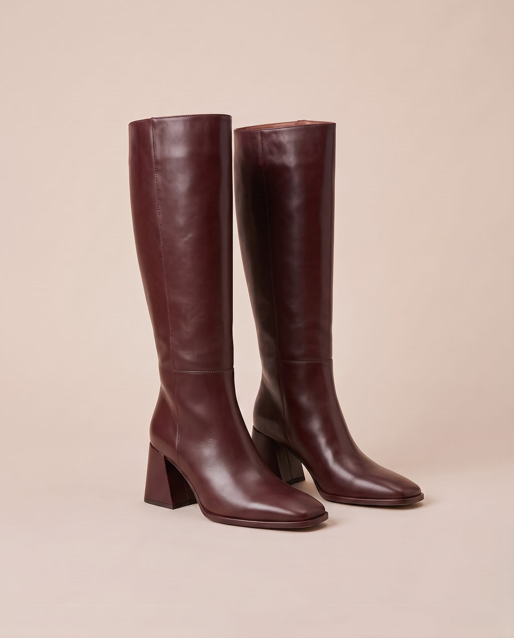 A pair of knee-high dark burgundy boots with block heels and pointed toes, displayed against a solid beige background.