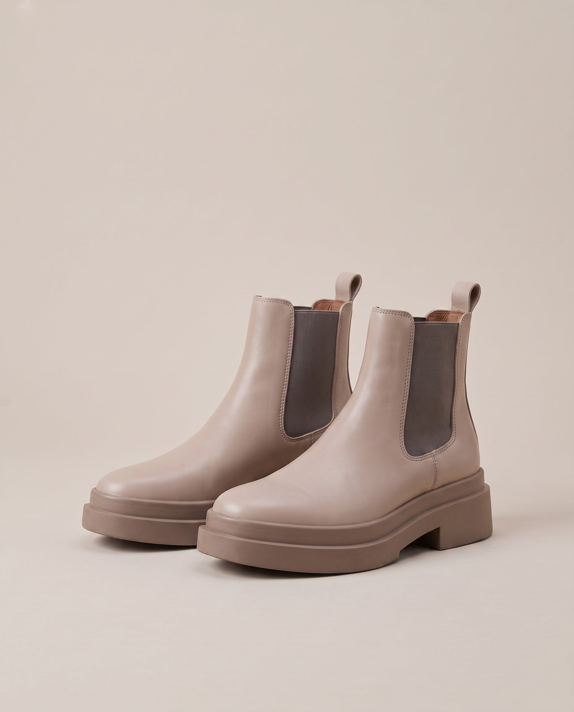 A pair of chunky platform Chelsea boots in a light taupe-beige color, featuring dark gray elastic side panels, photographed on a solid light beige background.