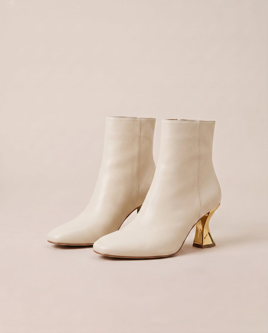 A pair of off-white ankle boots with pointed toes and sculptural gold block heels, displayed on a warm beige background.