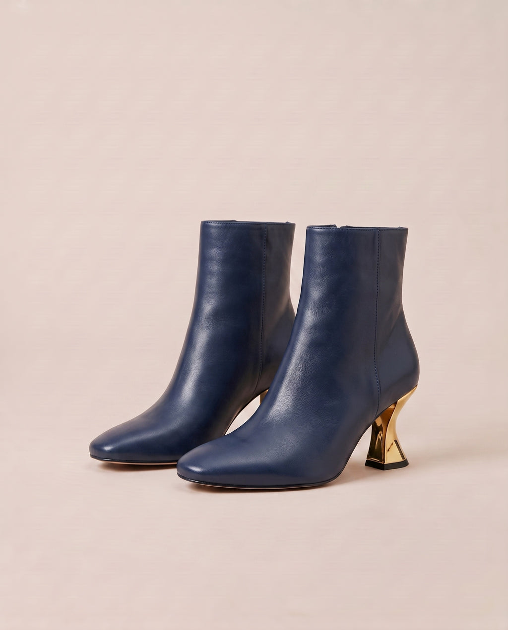 A pair of navy blue ankle boots with pointed toes and sculptural gold metal heels, displayed on a warm beige background.