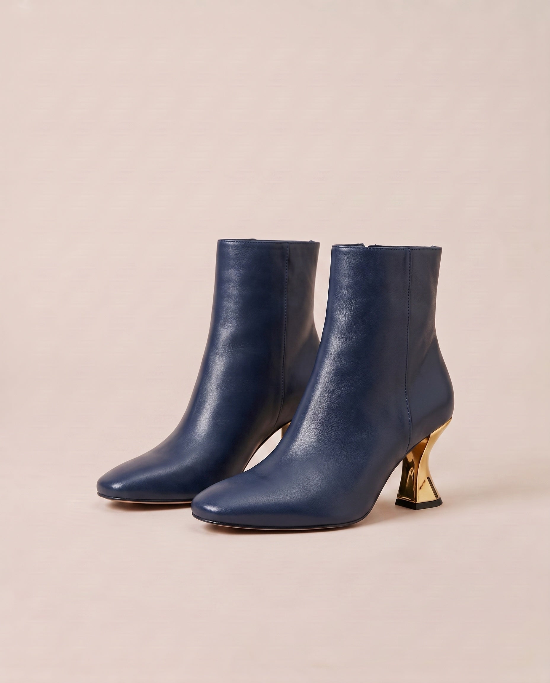 A pair of navy blue ankle boots with pointed toes and sculptural gold metal heels, displayed on a warm beige background.
