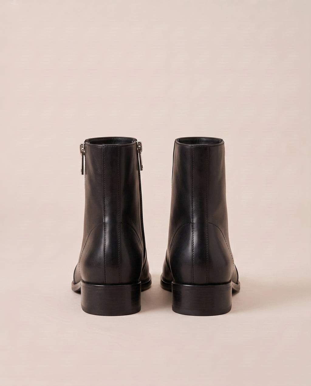 A pair of black ankle boots shown from the back, featuring side zippers and low block heels, set against a solid light beige background.