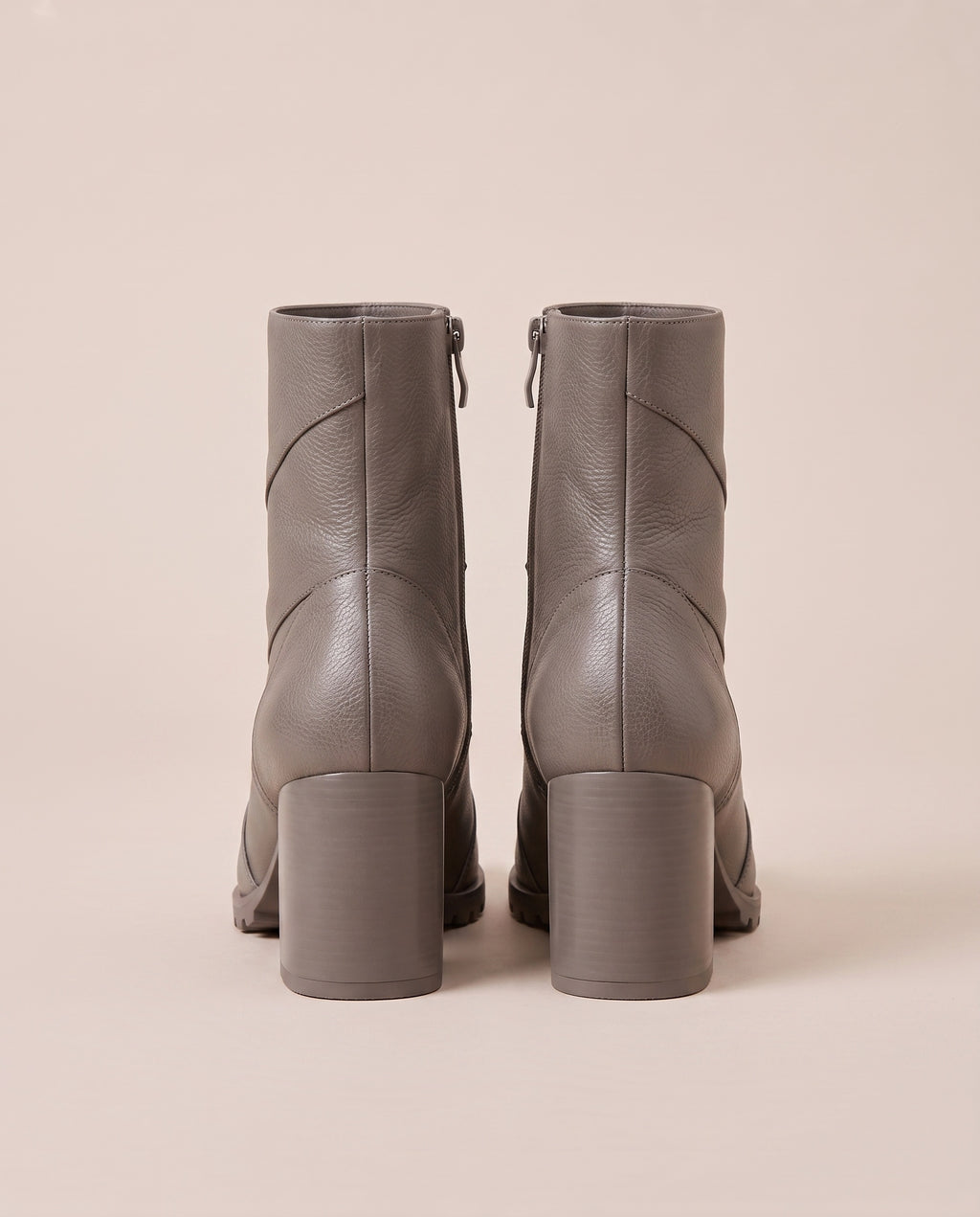 A pair of taupe gray ankle boots with block heels and side zippers, shown from the back against a solid beige background.