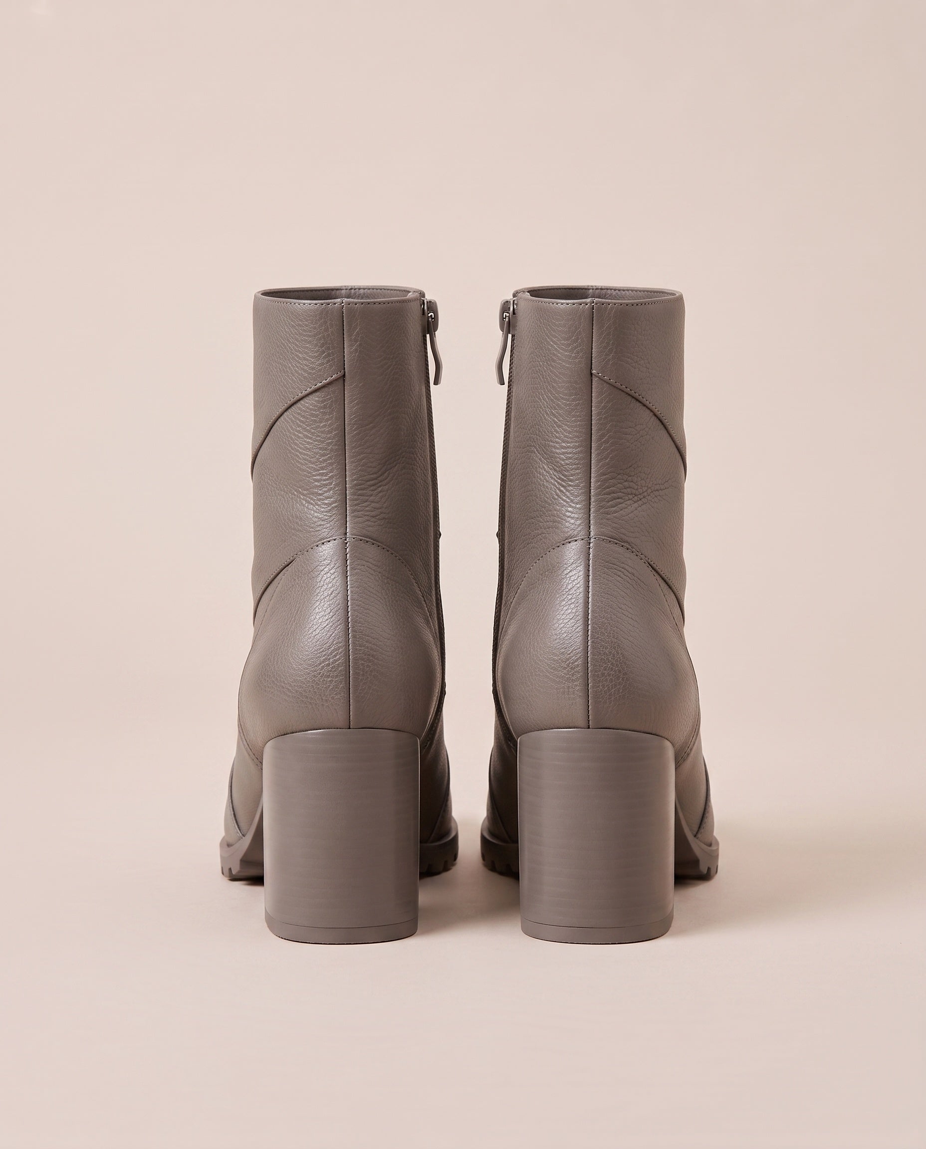 A pair of taupe gray ankle boots with block heels and side zippers, shown from the back against a solid beige background.