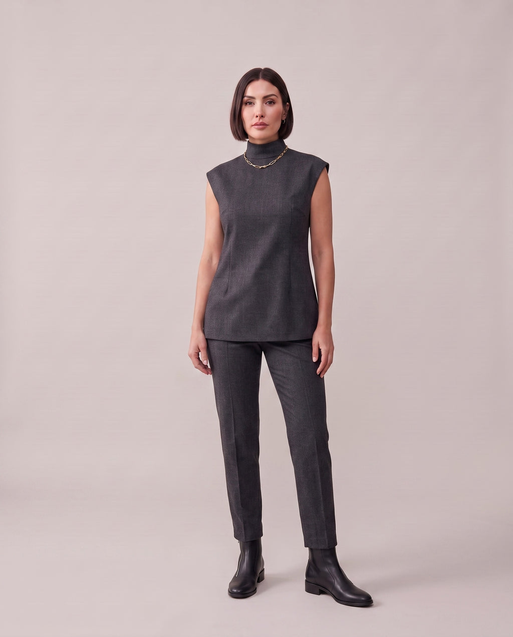 A woman modeling a dark gray sleeveless mock-neck top and matching dark gray trousers with black ankle boots, standing against a solid light beige studio background.