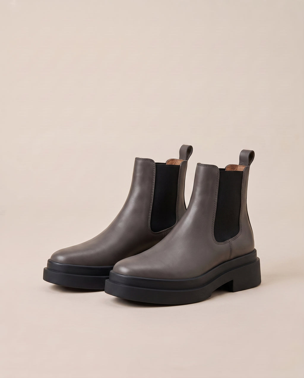 A pair of dark gray Chelsea boots with black elastic side panels and chunky black platform soles, displayed on a warm beige background.