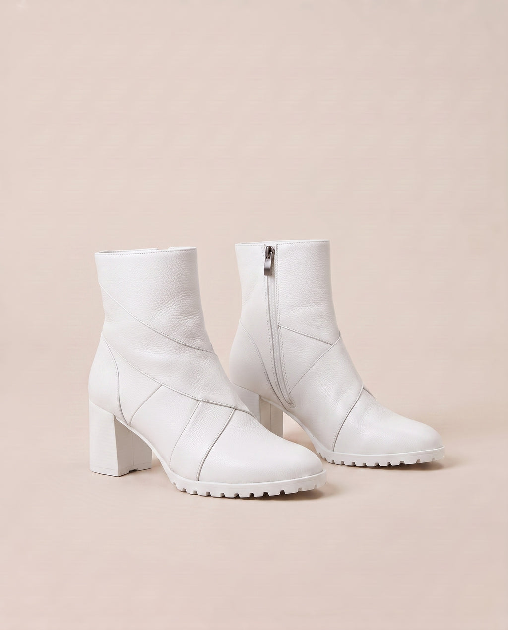 A pair of white ankle boots with a block heel, side zipper, and quilted geometric paneling, set against a solid pale beige background.
