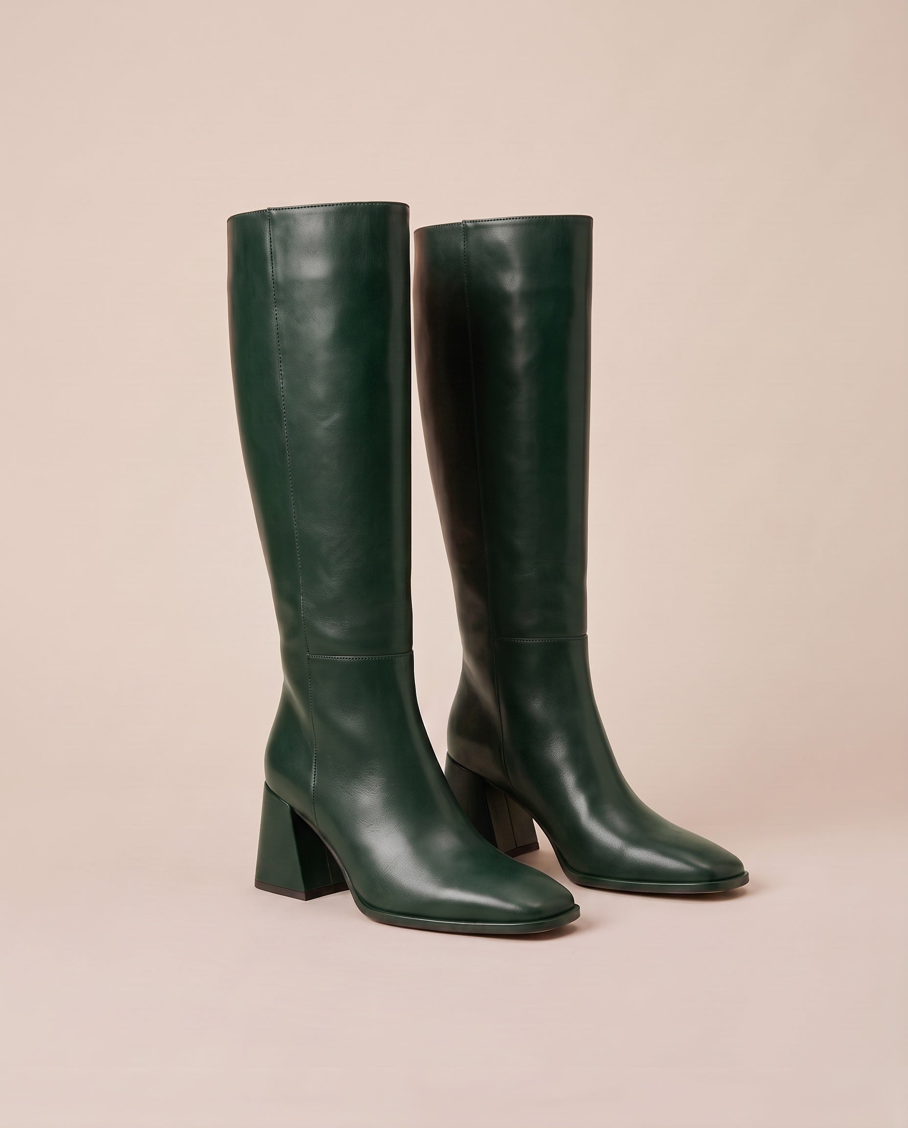 A pair of dark forest green knee-high boots with block heels and square toes, displayed against a solid pale pink background.