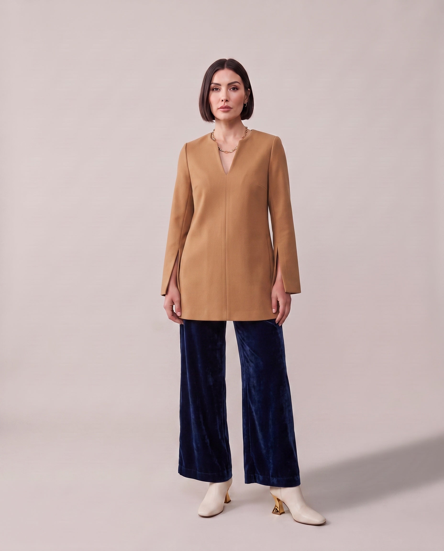 A woman modeling a camel-colored wrap-style jacket with a split neckline, paired with dark blue velvet wide-leg pants and white ankle boots, against a solid light beige studio background.
