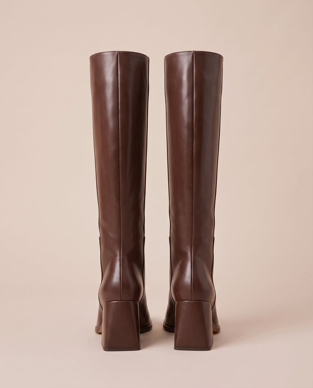 A pair of dark brown knee-high boots shown from the back, featuring a block heel and a straight shaft, set against a solid beige background.
