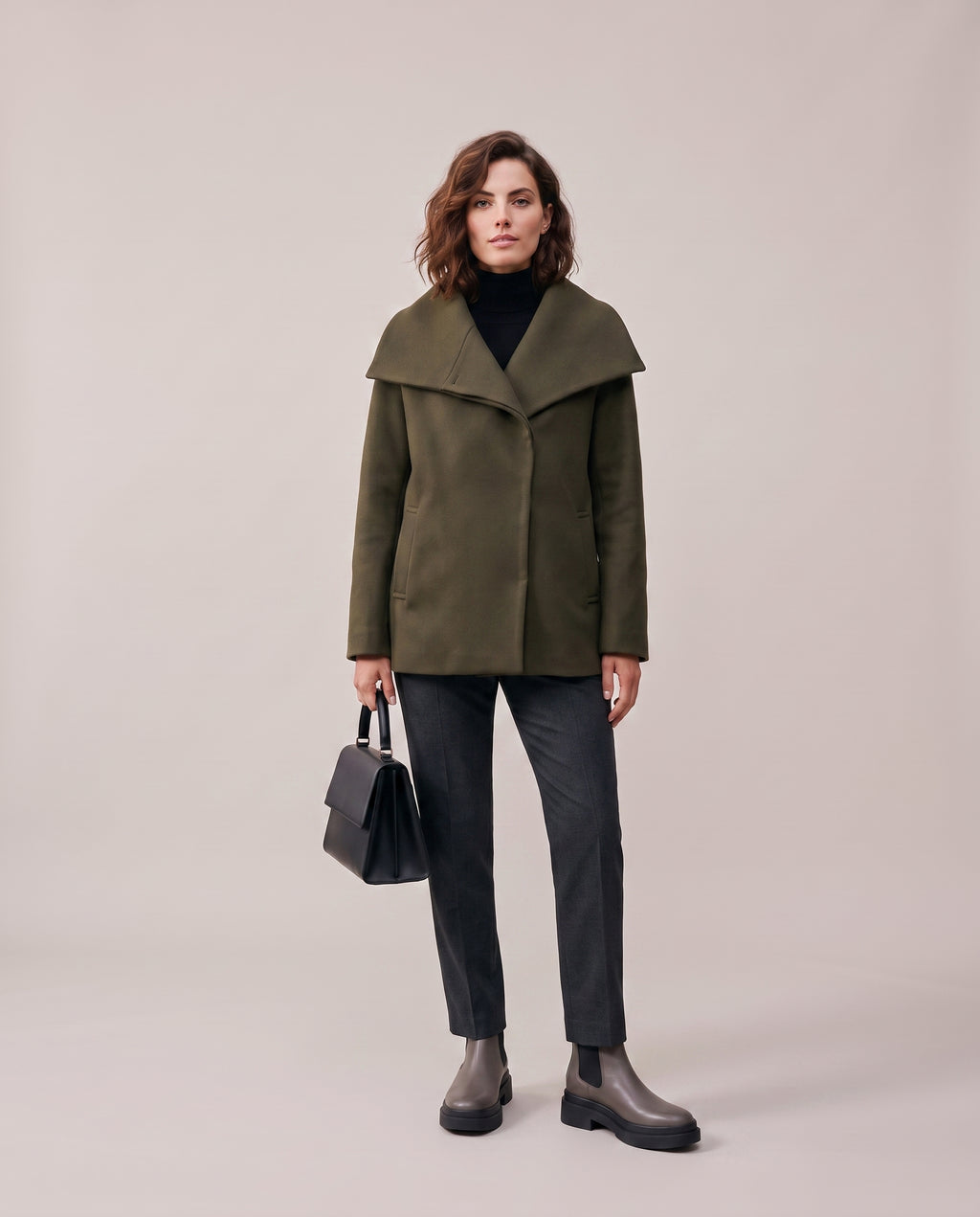 A woman stands against a plain light beige studio background wearing an olive green wide-collar coat over a black turtleneck, dark charcoal pants, dark gray Chelsea boots, and holding a black handbag.