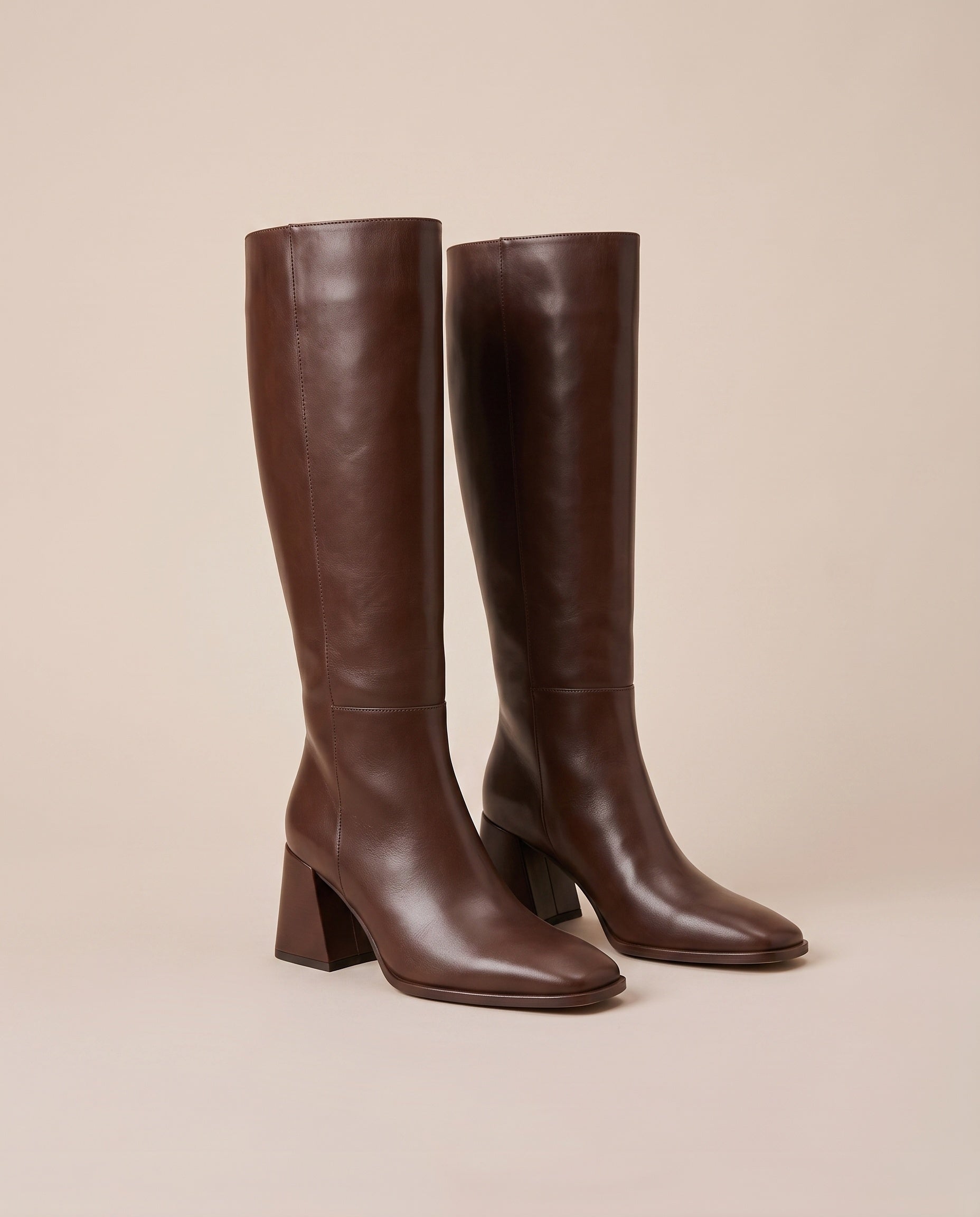 A pair of dark brown leather knee-high boots with block heels and square toes, displayed against a solid beige background.