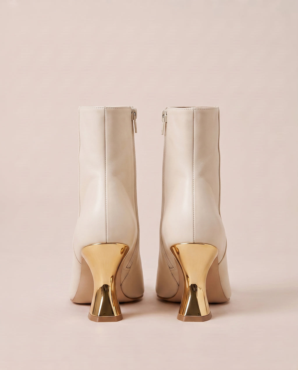 A pair of off-white ankle boots with distinctive metallic gold geometric block heels, shown from the back against a solid pale pink background.