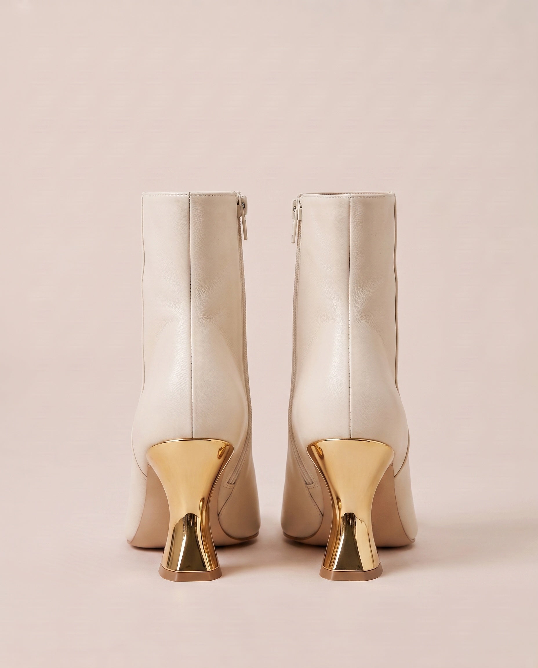 A pair of off-white ankle boots with distinctive metallic gold geometric block heels, shown from the back against a solid pale pink background.