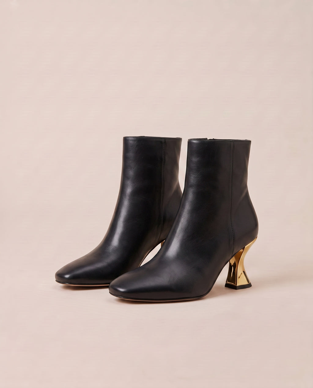 A pair of black ankle boots with a pointed toe and a distinctive metallic gold sculptural block heel, set against a solid light beige background.
