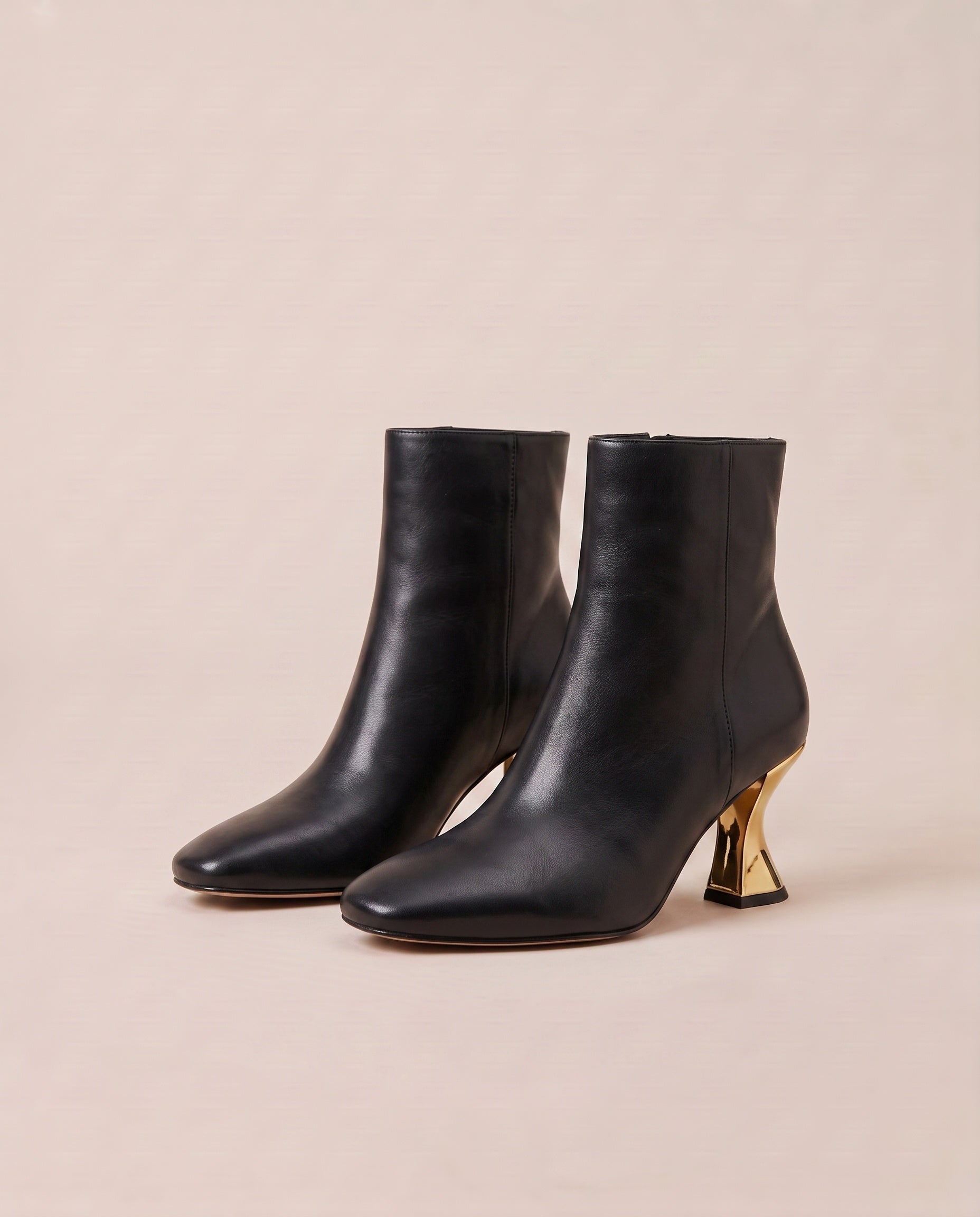 A pair of black ankle boots with a pointed toe and a distinctive metallic gold sculptural block heel, set against a solid light beige background.