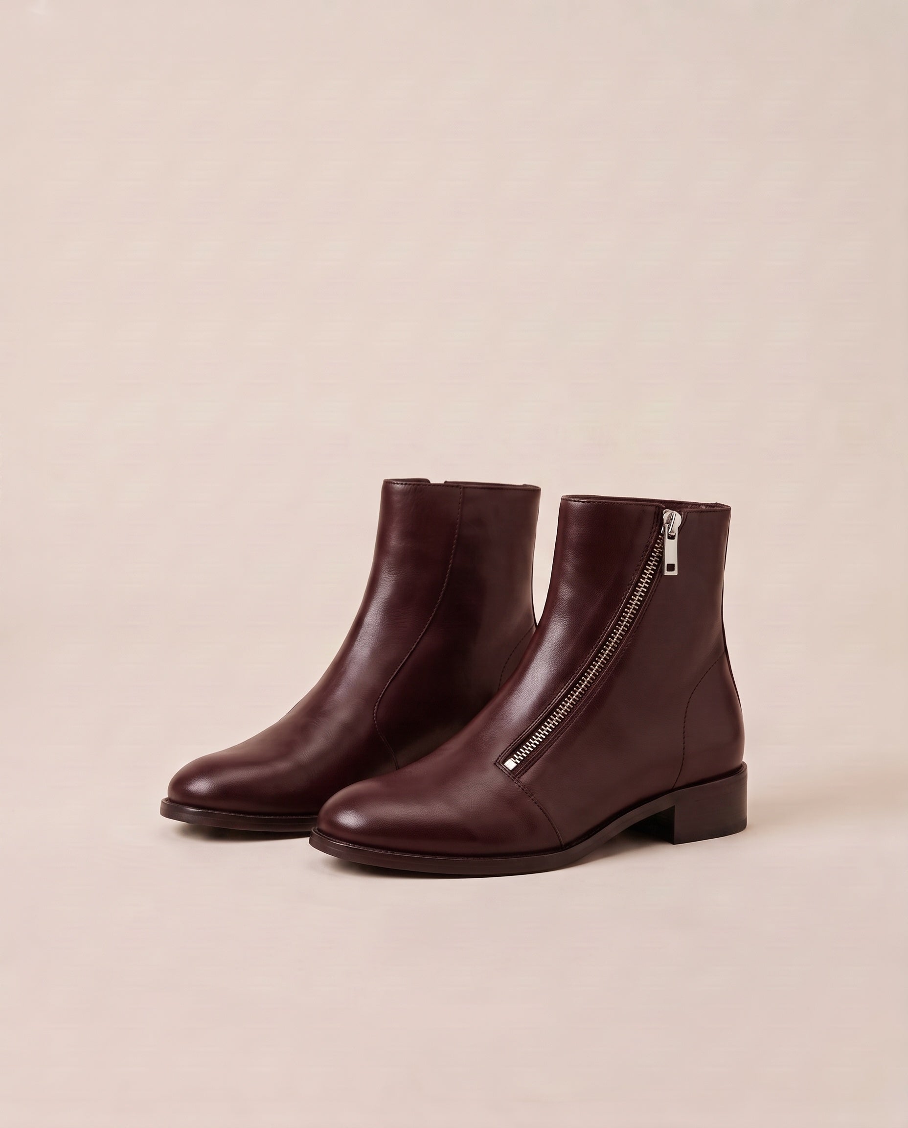 A pair of dark burgundy ankle boots with a side zipper and low block heel, displayed on a neutral beige background.