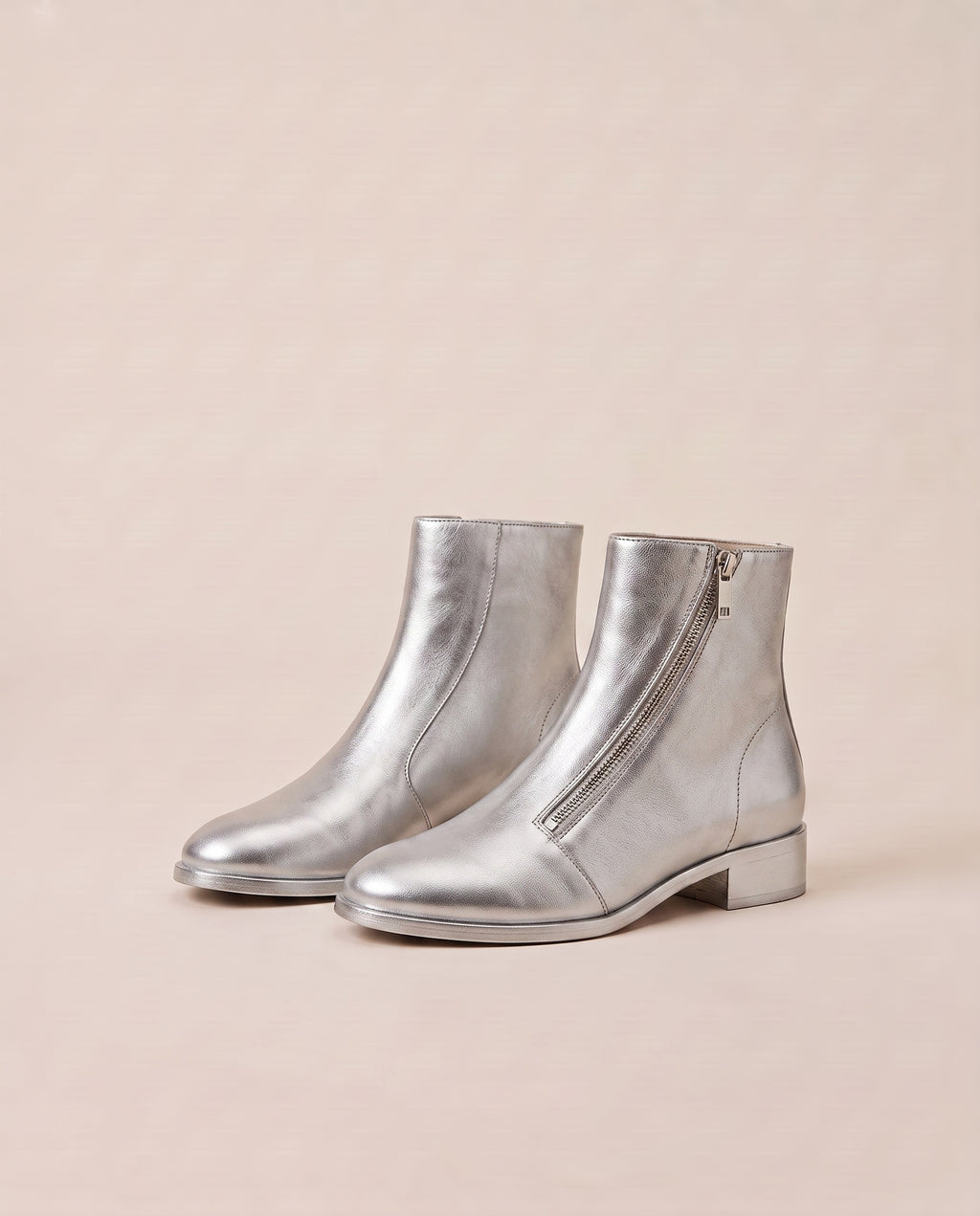 A pair of metallic silver ankle boots with low block heels and side zippers, displayed on a warm beige background.