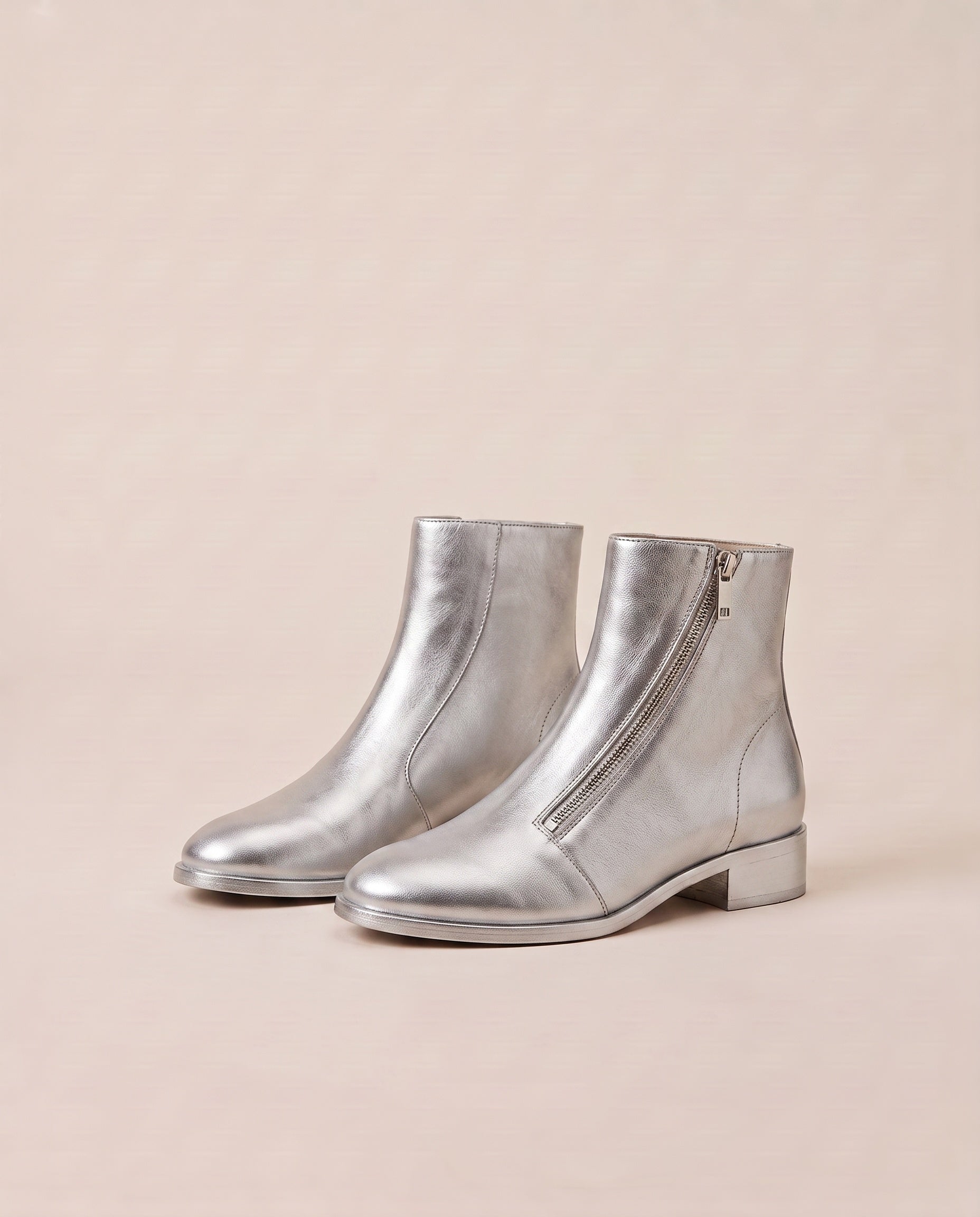 A pair of metallic silver ankle boots with low block heels and side zippers, displayed on a warm beige background.