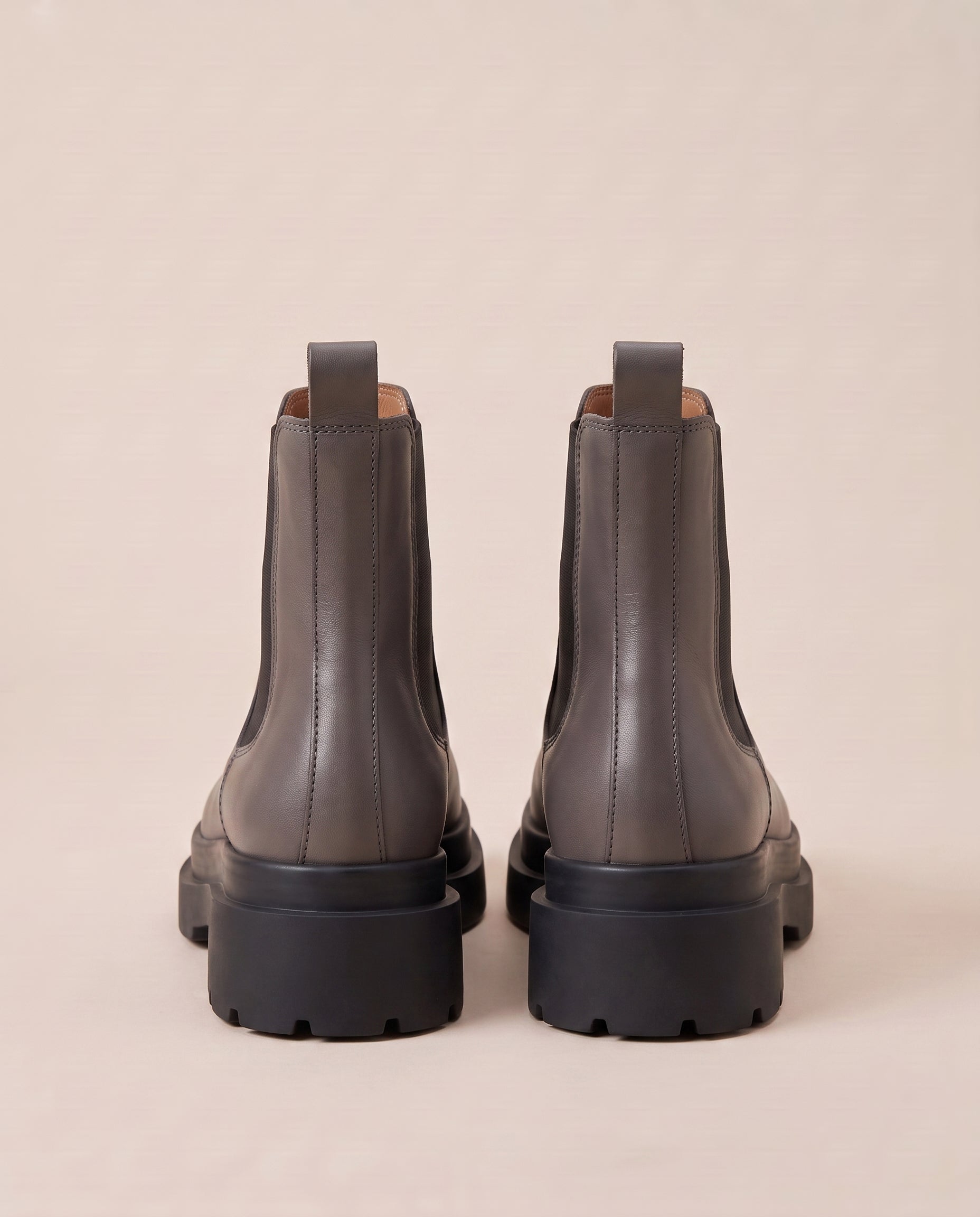 A pair of dark gray Chelsea boots with chunky black platform soles, shown from the back against a solid beige background.