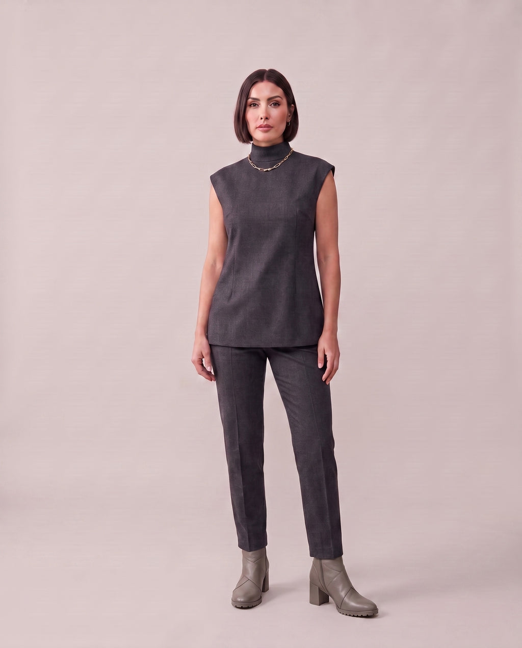 A woman modeling a dark gray sleeveless mock-neck top and matching dark gray trousers, paired with gray heeled ankle boots, against a solid light beige studio background.