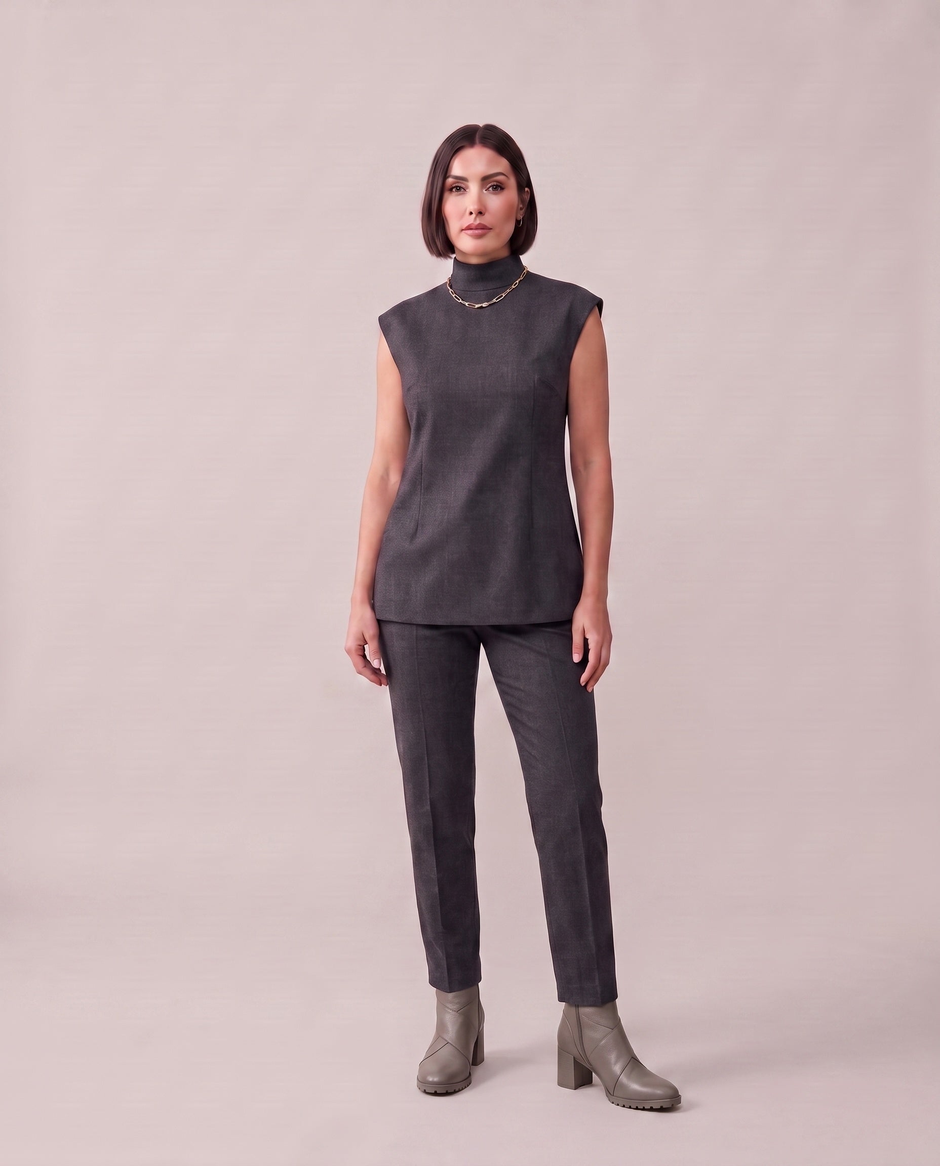 A woman modeling a dark gray sleeveless mock-neck top and matching dark gray trousers, paired with gray heeled ankle boots, against a solid light beige studio background.