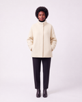 A model stands against a light gray studio background wearing a cream-colored, high-neck, oversized coat, black trousers, and black ankle boots.
