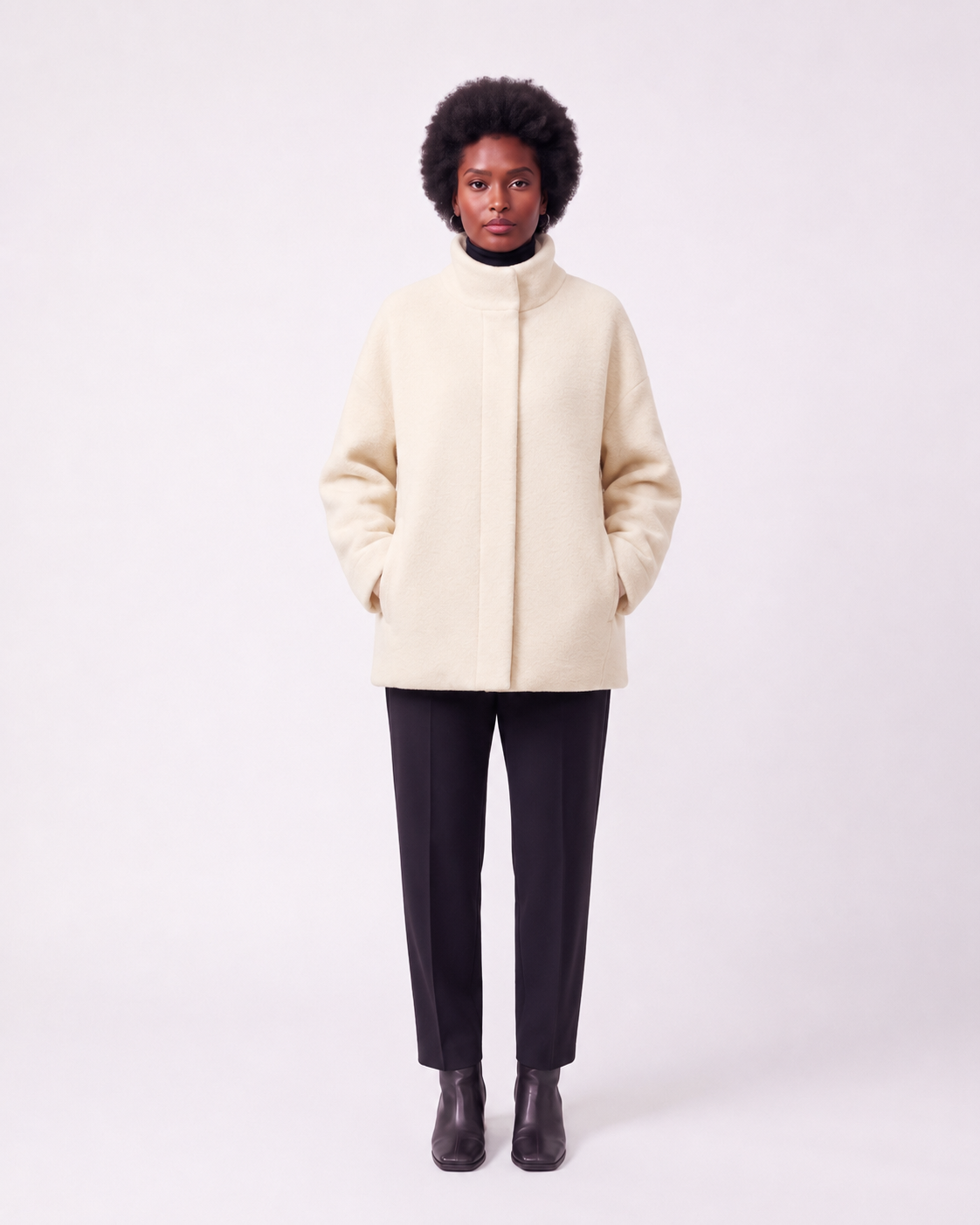 A model stands against a light gray studio background wearing a cream-colored, high-neck, oversized coat, black trousers, and black ankle boots.