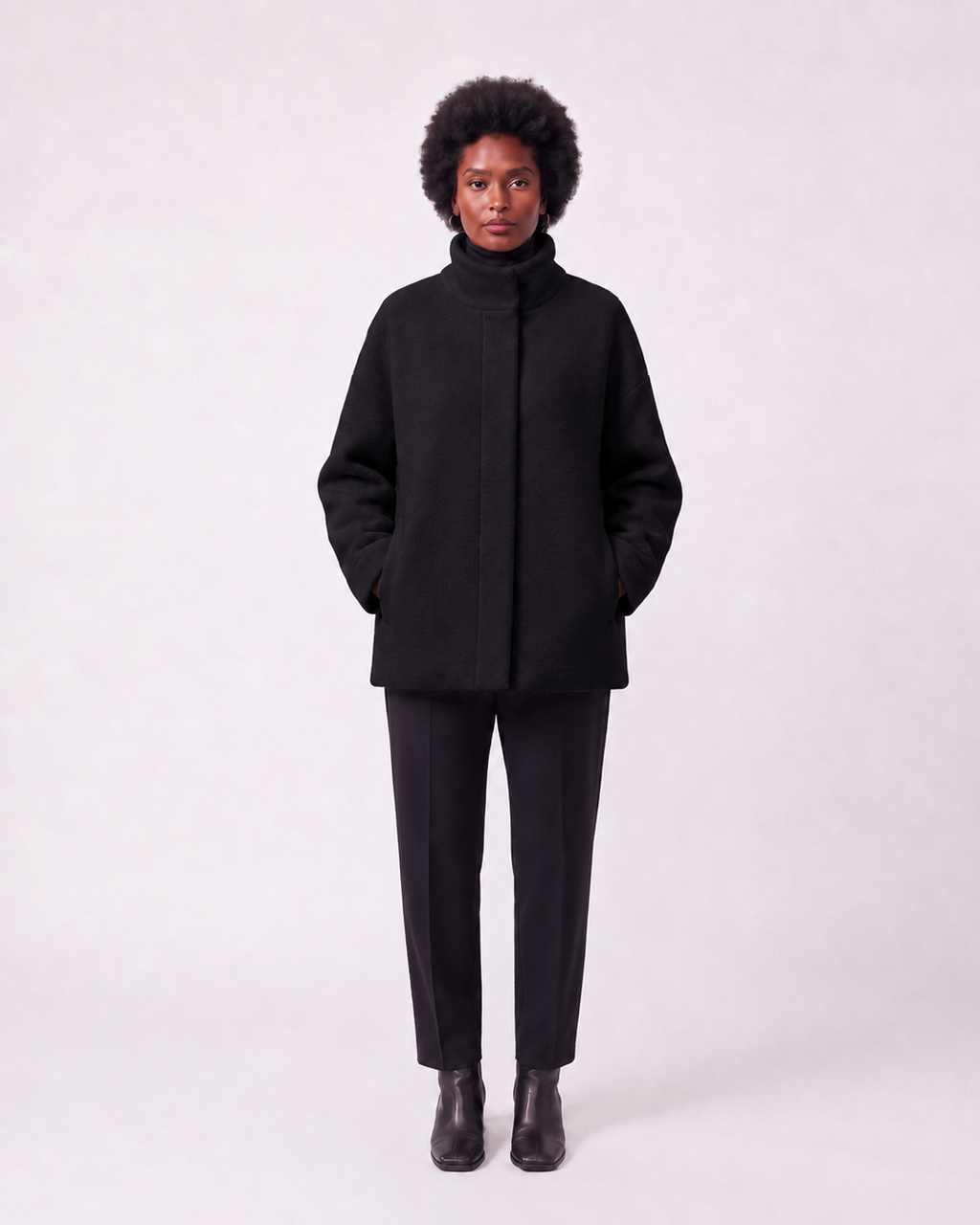 A model stands against a light gray studio background wearing a black high-neck coat, black trousers, and black ankle boots.