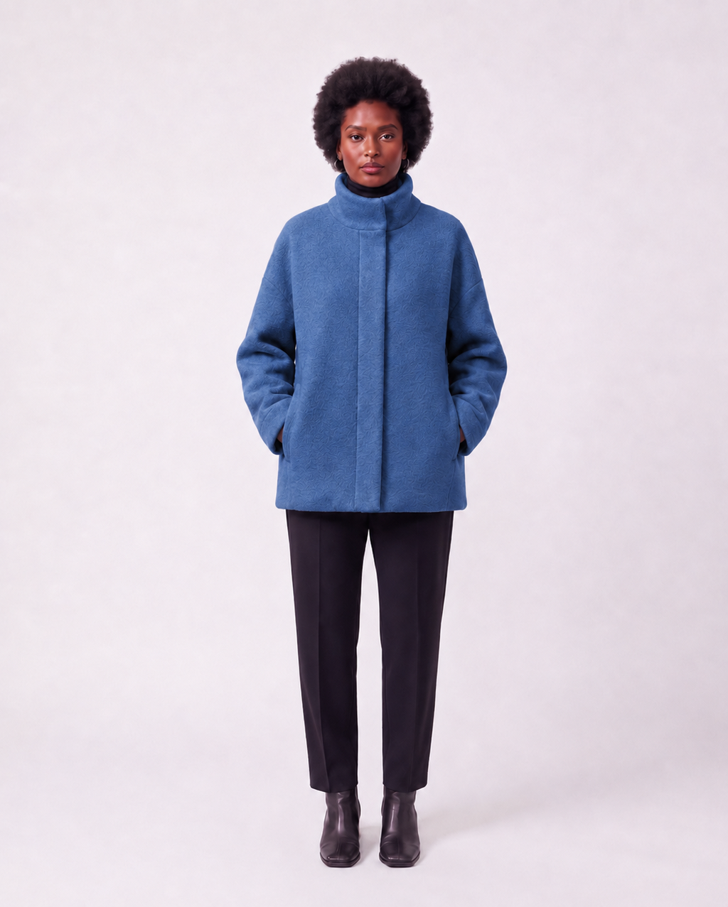 A model wearing a blue high-neck cropped coat, black trousers, and black boots, standing against a light gray studio background.