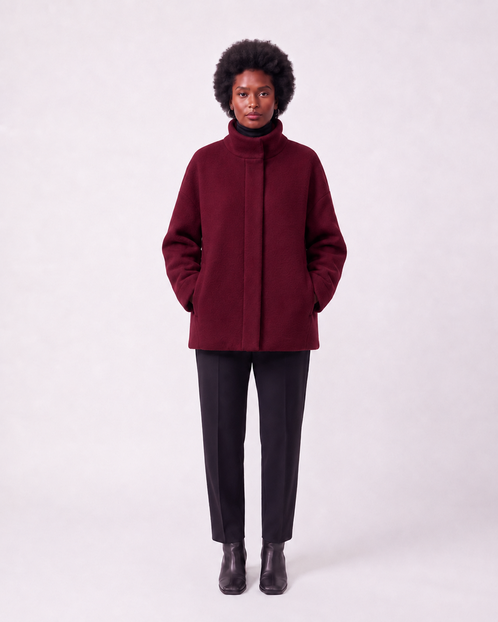 A model wearing a dark burgundy high-neck coat, black trousers, and black boots, standing against a light gray studio background.
