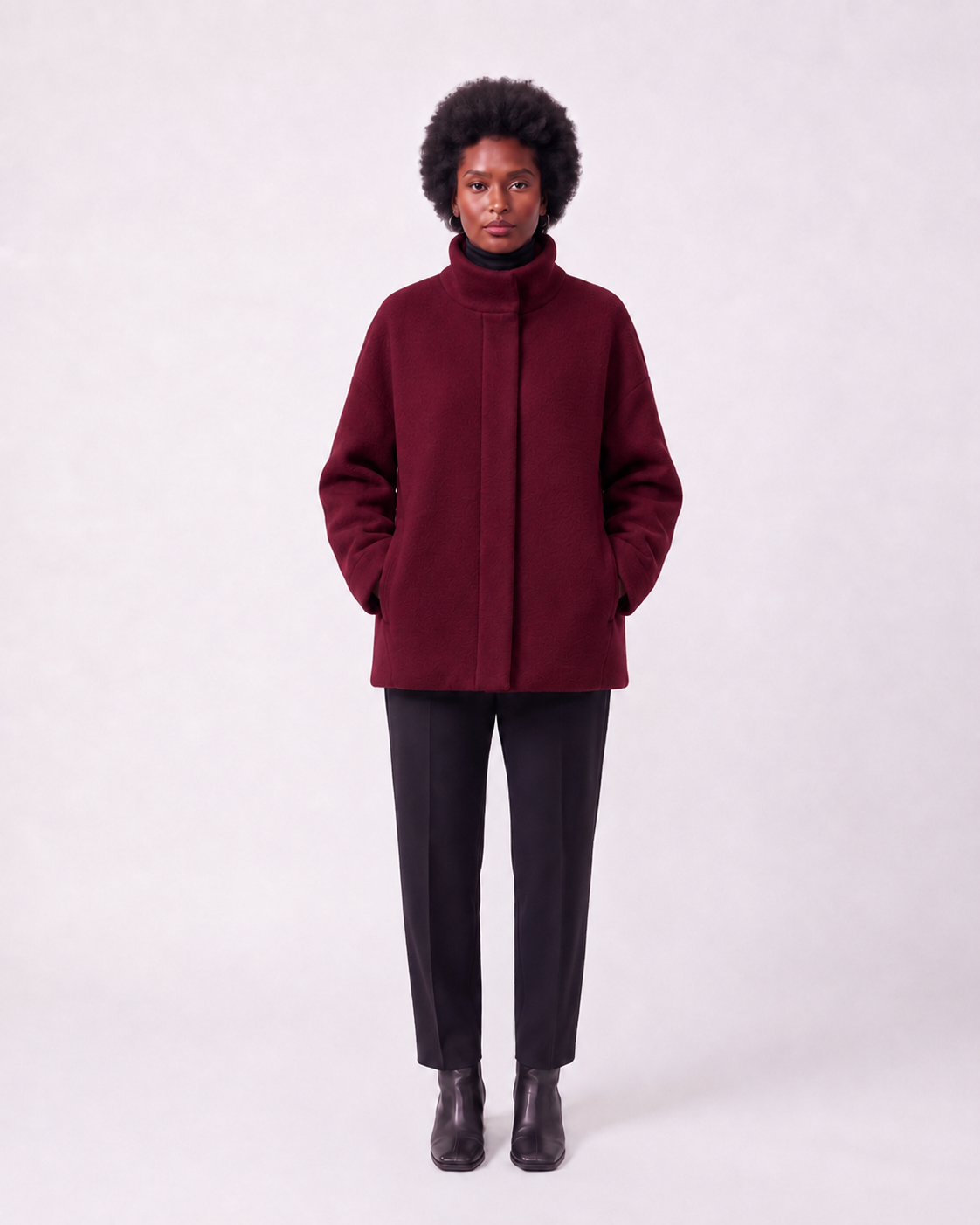 A model wearing a dark burgundy high-neck coat, black trousers, and black boots, standing against a light gray studio background.