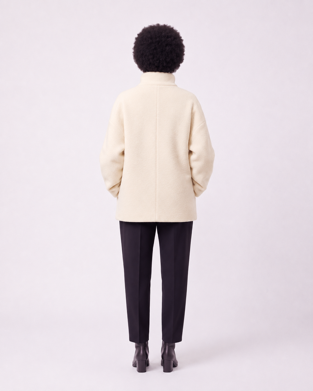 A person shown from the back wearing a cream-colored coat, dark trousers, and black shoes against a plain light gray studio background.