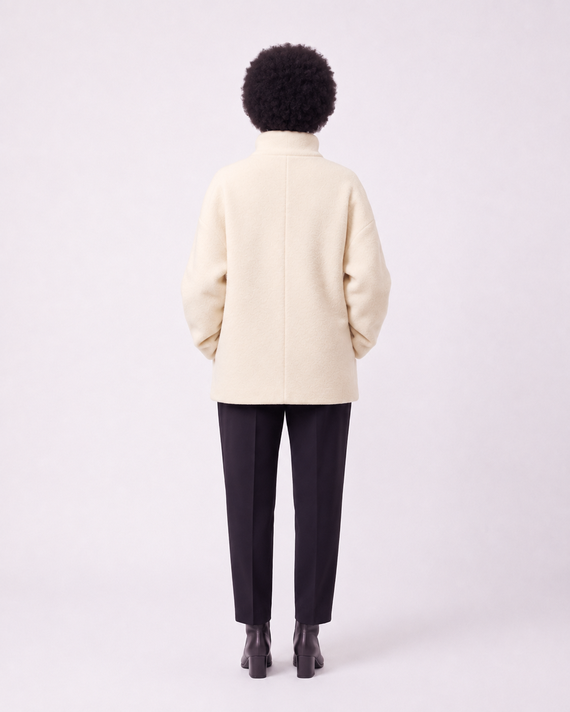 A person shown from the back wearing a cream-colored coat, dark trousers, and black shoes against a plain light gray studio background.