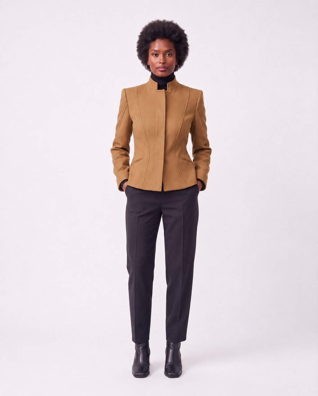 A model with an afro hairstyle stands against a light gray studio background wearing a structured camel-brown jacket with a high stand collar, paired with dark charcoal trousers and black ankle boots.