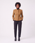 A model with an afro hairstyle stands against a light gray studio background wearing a structured camel-brown jacket with a high stand collar, paired with dark charcoal trousers and black ankle boots.