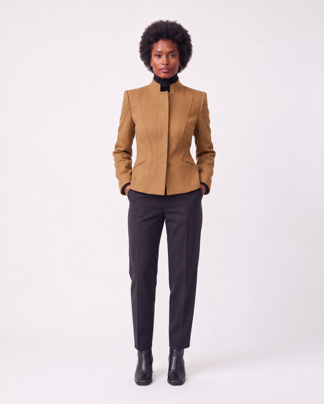 A model with an afro hairstyle stands against a light gray studio background wearing a structured camel-brown jacket with a high stand collar, paired with dark charcoal trousers and black ankle boots.
