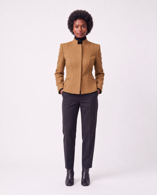 A model with an afro hairstyle stands against a light gray studio background wearing a structured camel-brown jacket with a high stand collar, paired with dark charcoal trousers and black ankle boots.