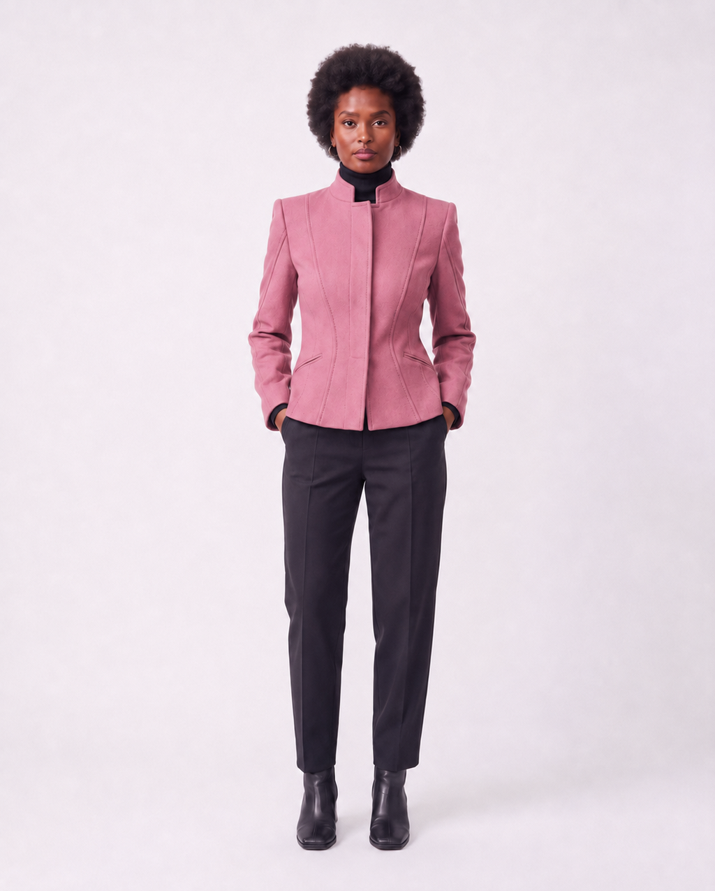 A model stands against a light gray studio background wearing a structured pink blazer with a high stand collar, paired with dark charcoal trousers and black ankle boots.