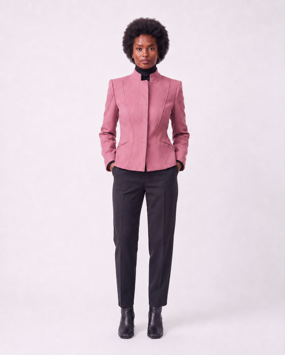 A model stands against a light gray studio background wearing a structured pink blazer with a high stand collar, paired with dark charcoal trousers and black ankle boots.